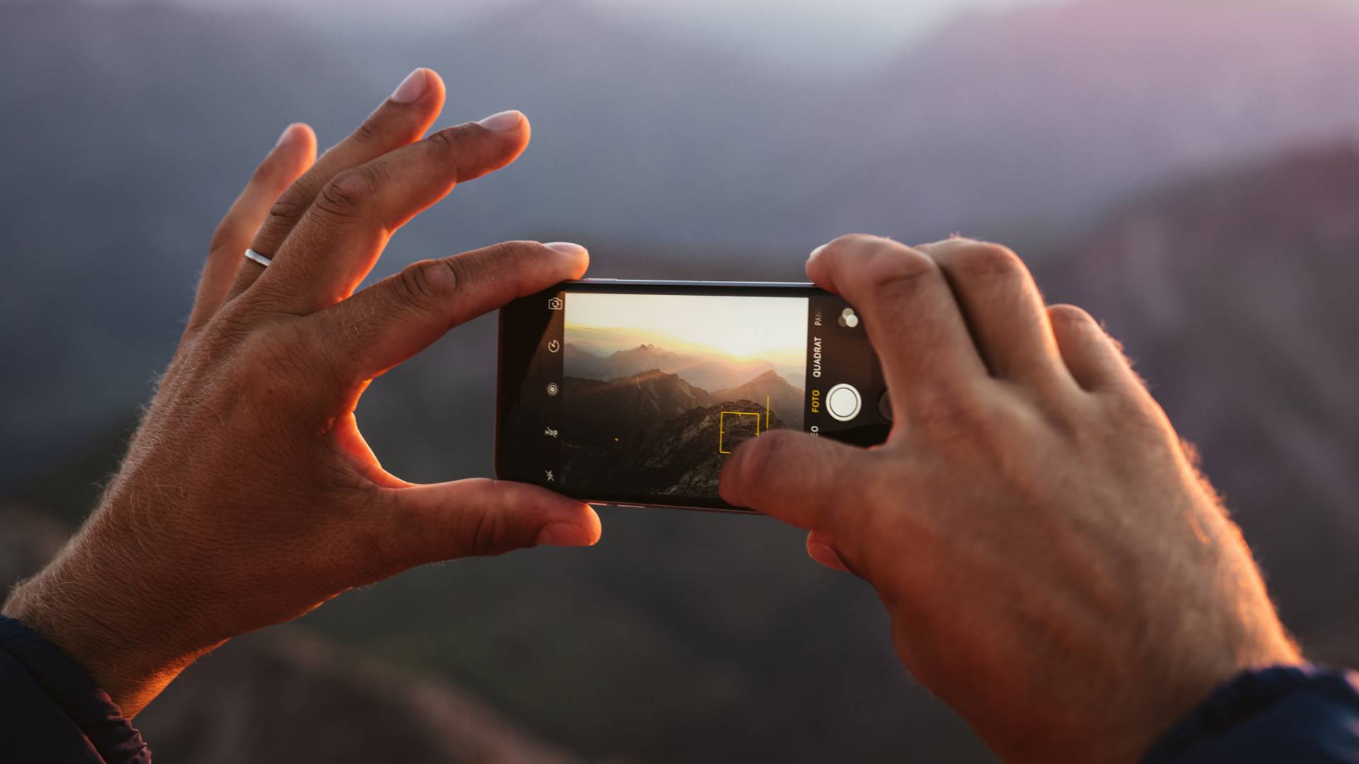 Hands taking a photo of a mountain landscape with a smartphone at sunset.